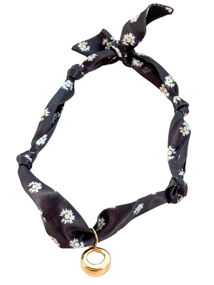 The Cuneo Scarf Necklace by Kinsey Designs features black floral fabric, a gold vintage-inspired circular pendant, tied ends, and an adjustable design, displayed on a white background.