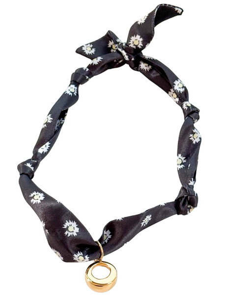 The Cuneo Scarf Necklace by Kinsey Designs features black floral fabric, a gold vintage-inspired circular pendant, tied ends, and an adjustable design, displayed on a white background.