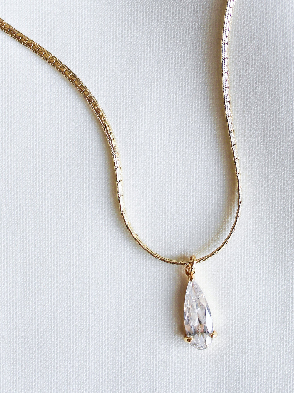 The Charlotte Drop Necklace by Kinsey Designs features a gold filled teardrop design with a clear cubic zirconia pendant, elegantly showcased on a white fabric background.