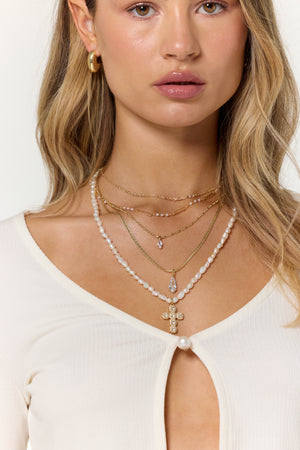 A woman wears the Kinsey Designs Charlotte Drop Necklace, layered with gold-filled and pearl necklaces including a cross pendant, paired with a cream top featuring a pearl button.