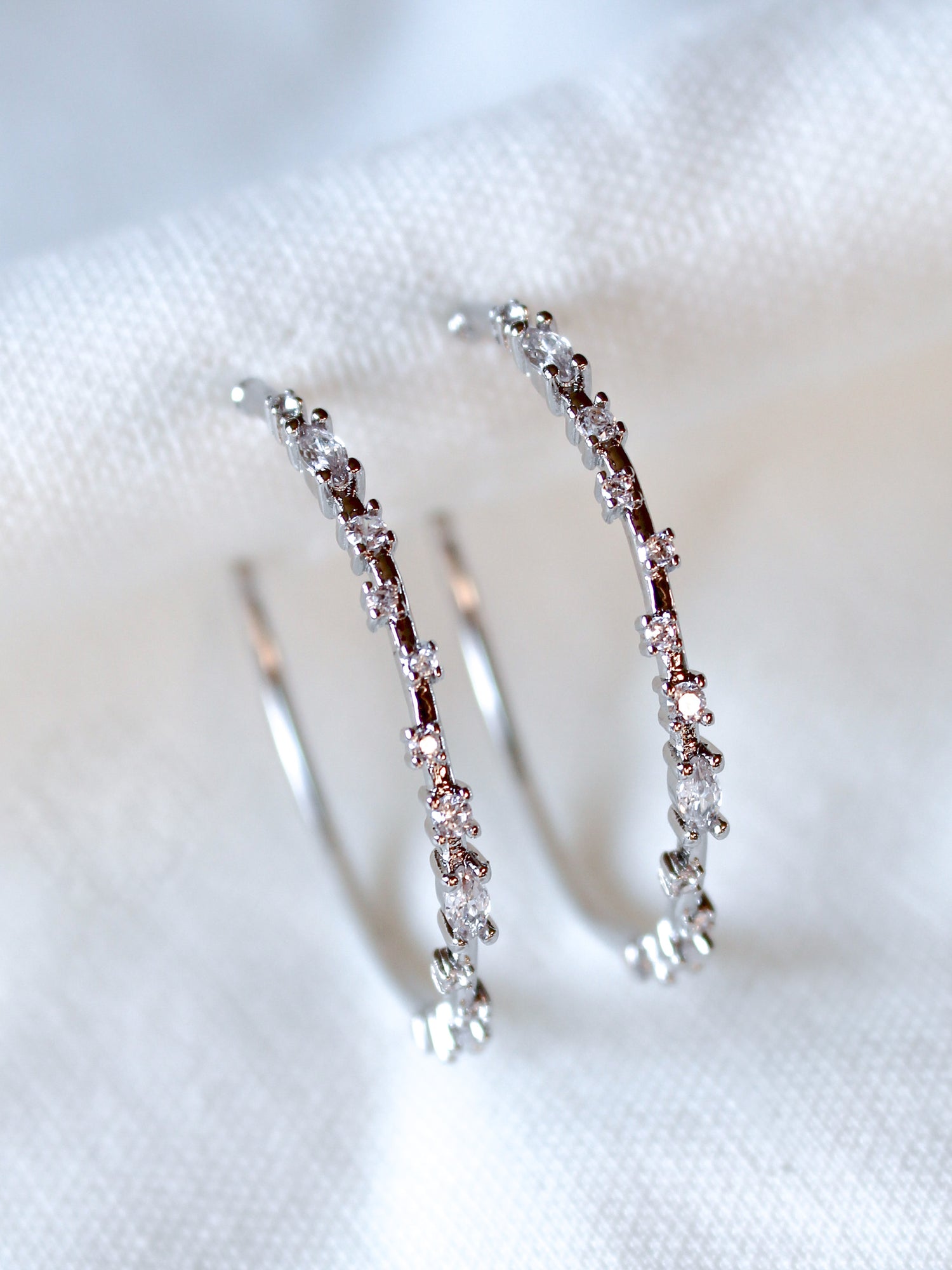 The Chase Hoop Large earrings by Kinsey Designs are silver hoops embellished with sparkling cubic zirconia, shown on a white fabric background.