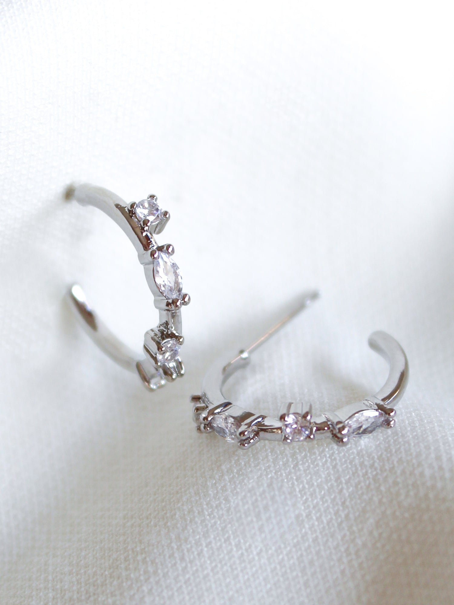 The Kinsey Designs Chase Hoop Mini Silver earrings feature two 0.5" diameter silver hoops adorned with small clear cubic zirconia gemstones, shown resting on a white textured fabric.
