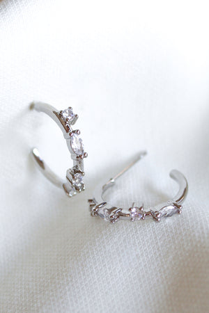 The Kinsey Designs Chase Hoop Mini Silver earrings feature two 0.5" diameter silver hoops adorned with small clear cubic zirconia gemstones, shown resting on a white textured fabric.