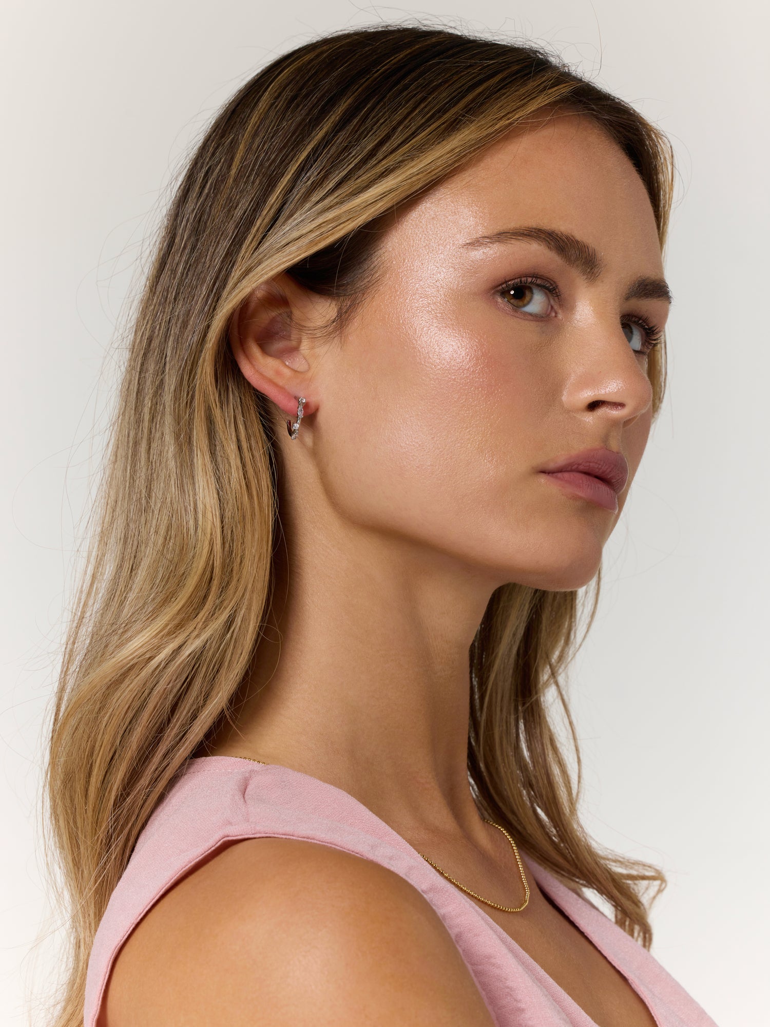 A woman with long, light brown hair wearing a pink top, gold necklace, and Kinsey Designs’ Chase Hoop Mini Silver earrings (.5" diameter), crafted from rhodium plated brass with cubic zirconia accents, looking sideways.