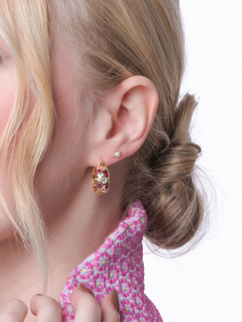 A woman with blonde hair in a bun wears Kinsey Designs’ Ruby Claire Hoops, stud earrings, a pink textured top, and subtle crystal accents for extra sparkle.