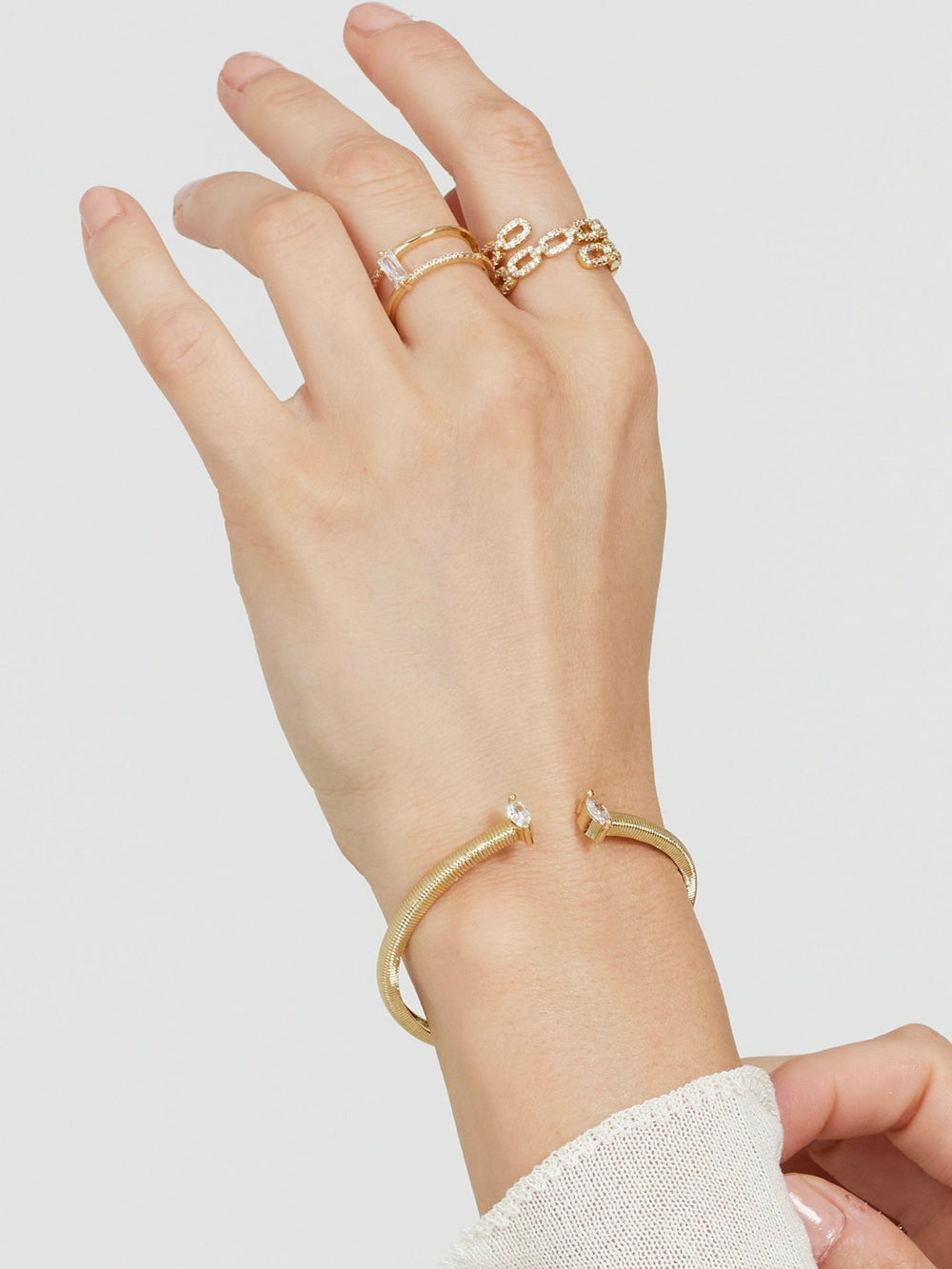 A hand wears Kinsey Designs' Cora Ring—an open gold ring with a baguette CZ stone—and a gold cuff bracelet with crystal ends, set against a plain light background.