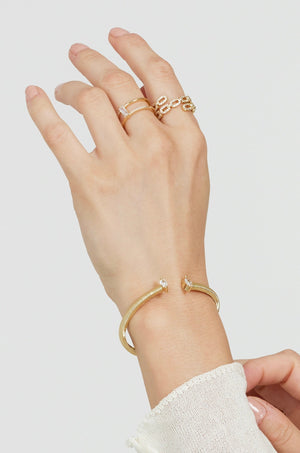 A hand wears Kinsey Designs' Cora Ring—an open gold ring with a baguette CZ stone—and a gold cuff bracelet with crystal ends, set against a plain light background.