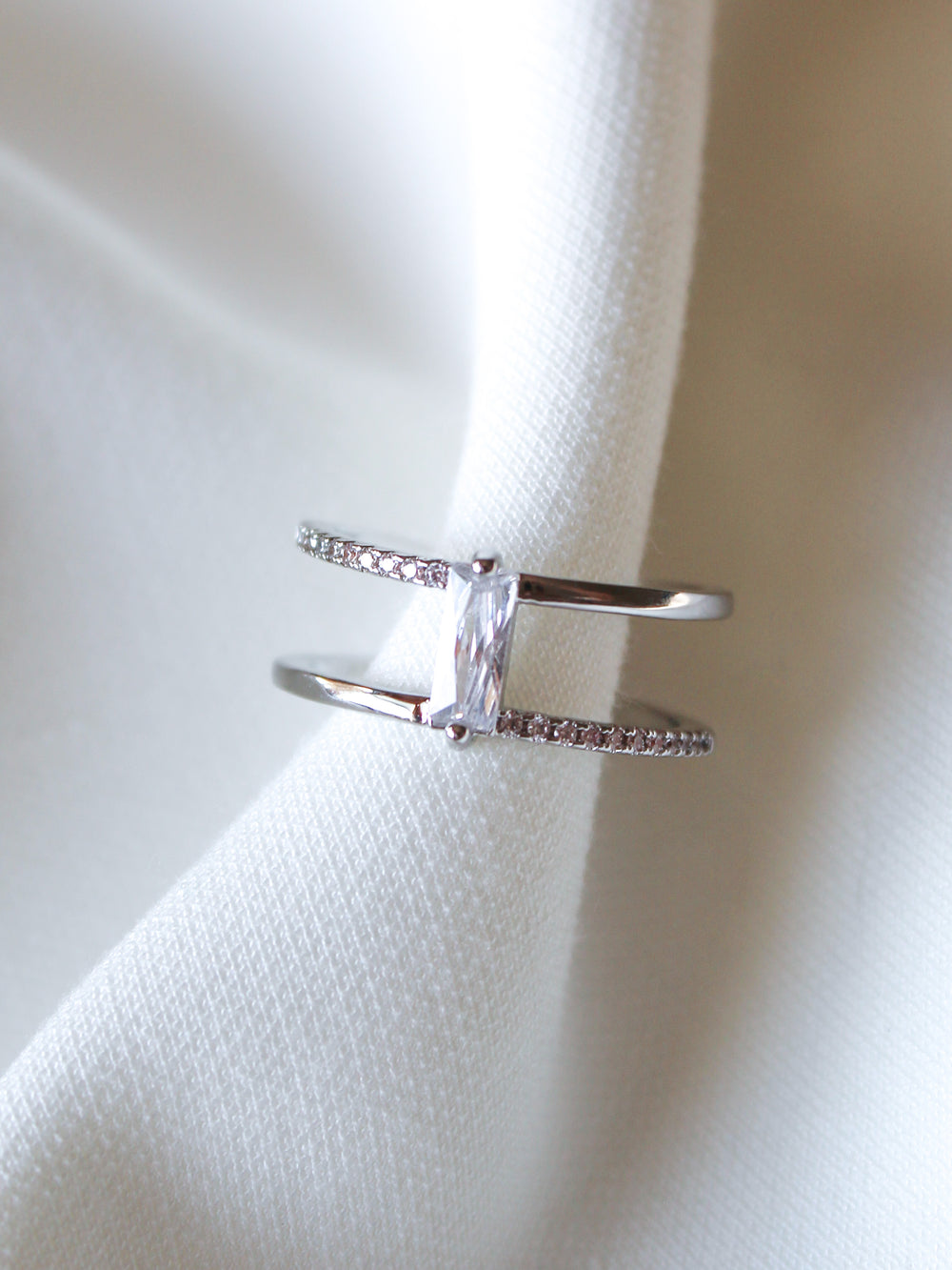 The Cora Ring by Kinsey Designs is an elegant open ring with a rectangular baguette CZ stone set between two bands adorned with small crystals, shown on white fabric.