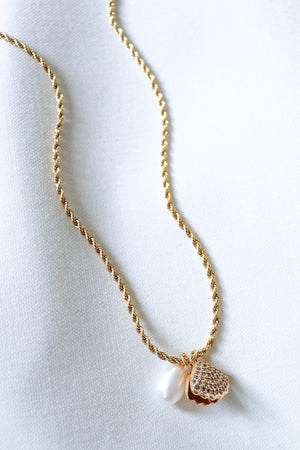 The Cove Pearl Necklace by Kinsey Designs Archives features a white pearl and textured gold heart pendant, elegantly displayed on a white fabric background.