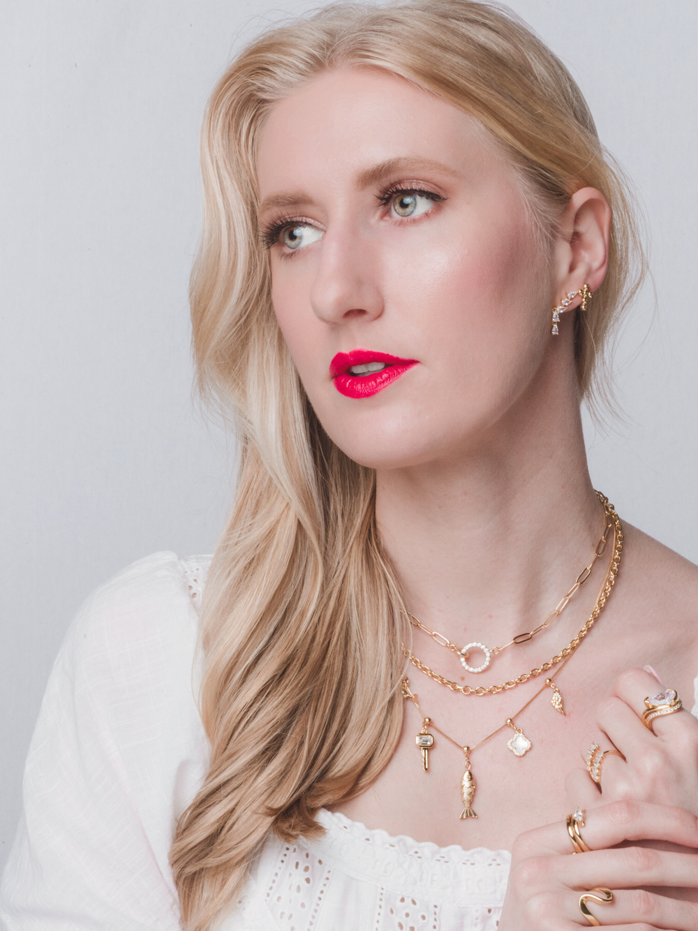 Blonde woman in a white top and bright pink lipstick looks to the side, wearing Kinsey Designs’ Cyrus Interlocking Circle Necklace in layered gold jewelry.