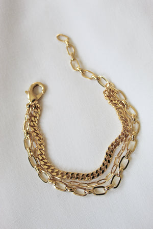The Declan Bracelet from Kinsey Designs features three unique gold chains in 6" layers, shown on a white fabric background.