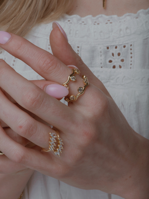 A woman's hand with pink nails showcases the Dove Stacking Ring Set by Kinsey Designs, featuring gemstone-adorned gold circle rings, against a white lace top.