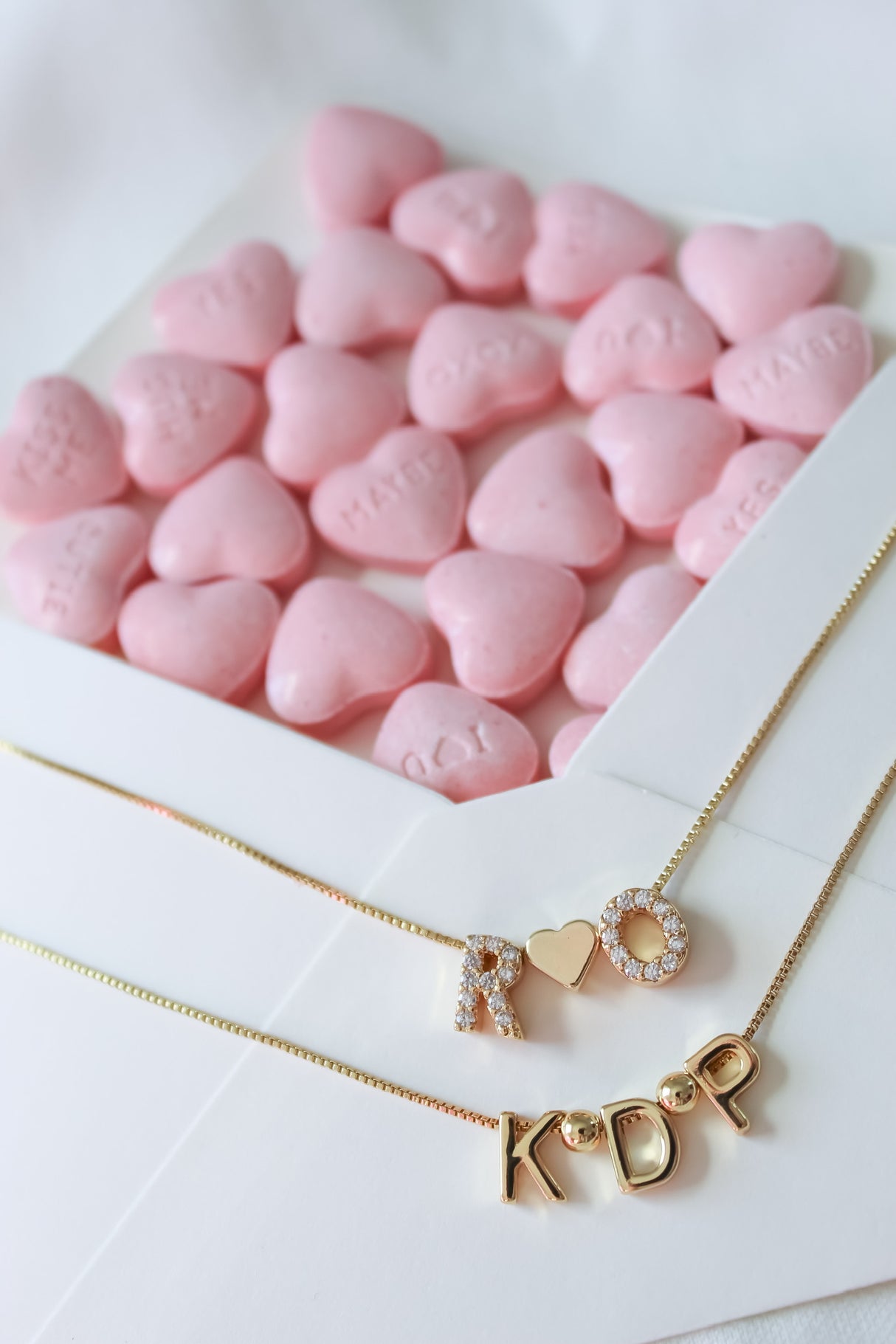 Gold necklaces with initials "R," "K," "D," and "P" placed in front of pink heart-shaped candies in an open white envelope, featuring the elegant Heart Spacer Slide from Kinsey Charm Bar.