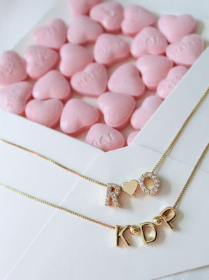 Two Kinsey Designs gold necklaces with letter charms and a Round Spacer Slide, displayed on pink heart candies and a white paper background.