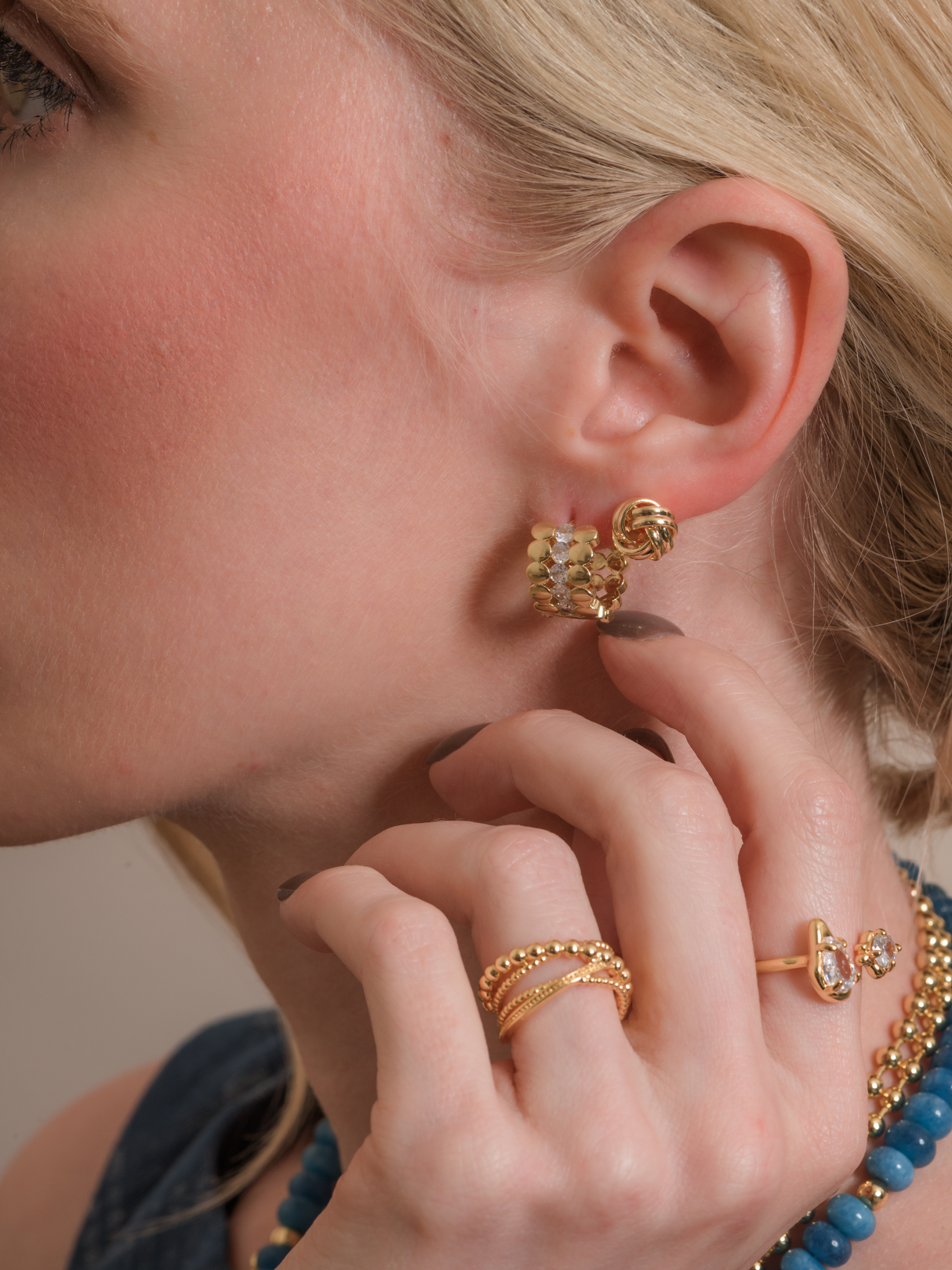 Close-up of a woman touching her Ellis Huggie Earring by Kinsey Designs, paired with gold rings and a blue beaded necklace.