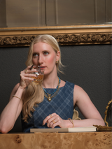 A woman in a blue dress, wearing the Ellis Huggie Earring by Kinsey Designs, sits at a table sipping from a glass, appearing thoughtful.