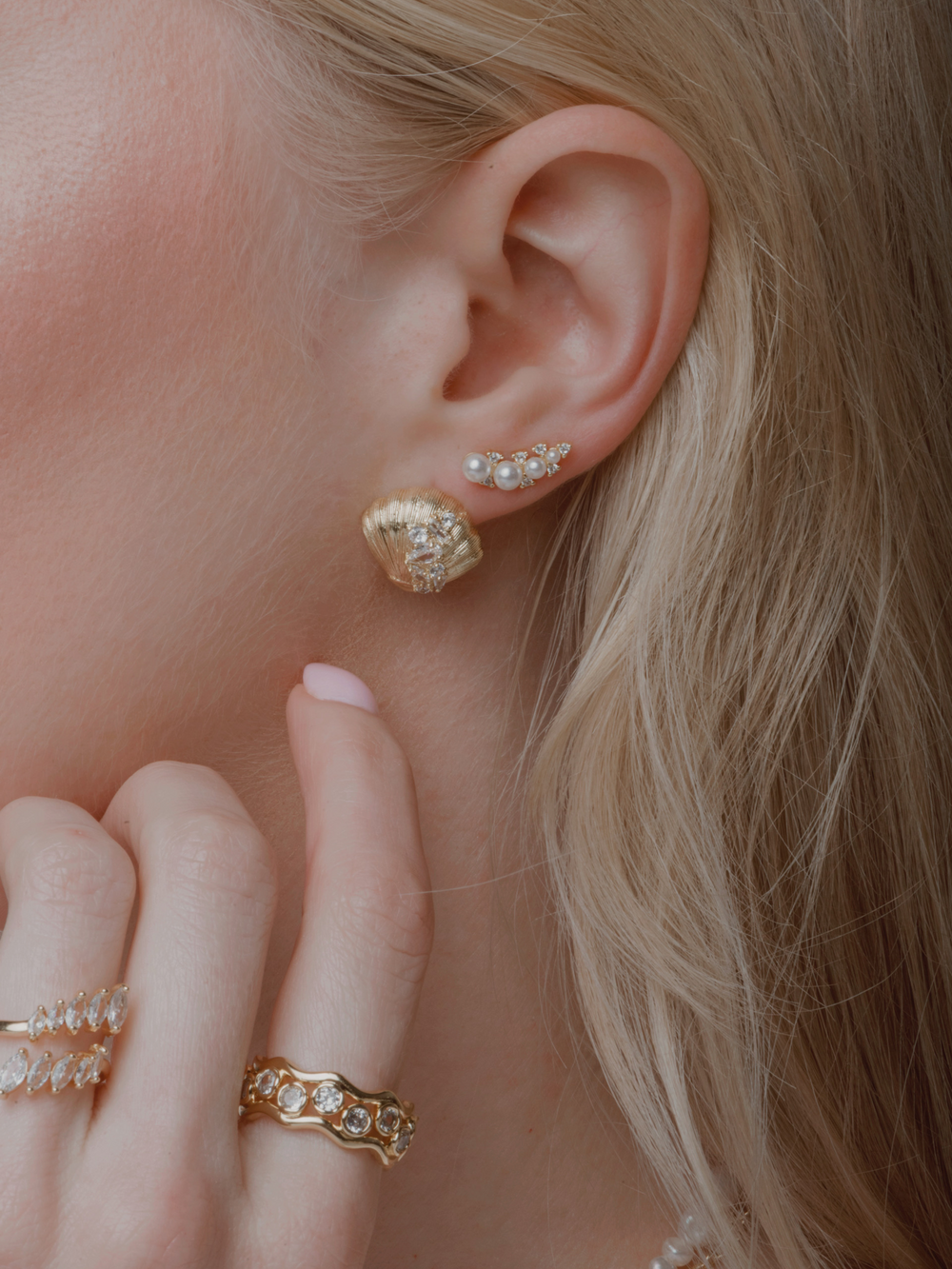 Close-up of a woman's ear wearing Kinsey Designs’ Lucian Seashell Post Earring, along with gold and pearl earrings, and her hand adorned with gold and diamond rings.