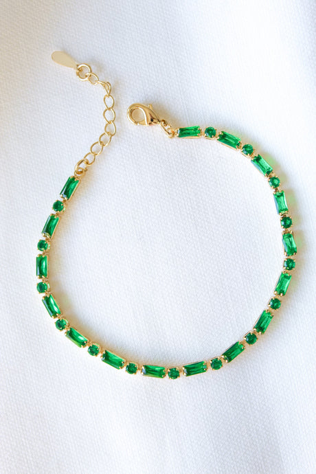 The Ella Bracelet by Kinsey Designs Archives features gold plating and rectangular green gemstones, shown on a white fabric. This adjustable piece adds everyday elegance to any look.