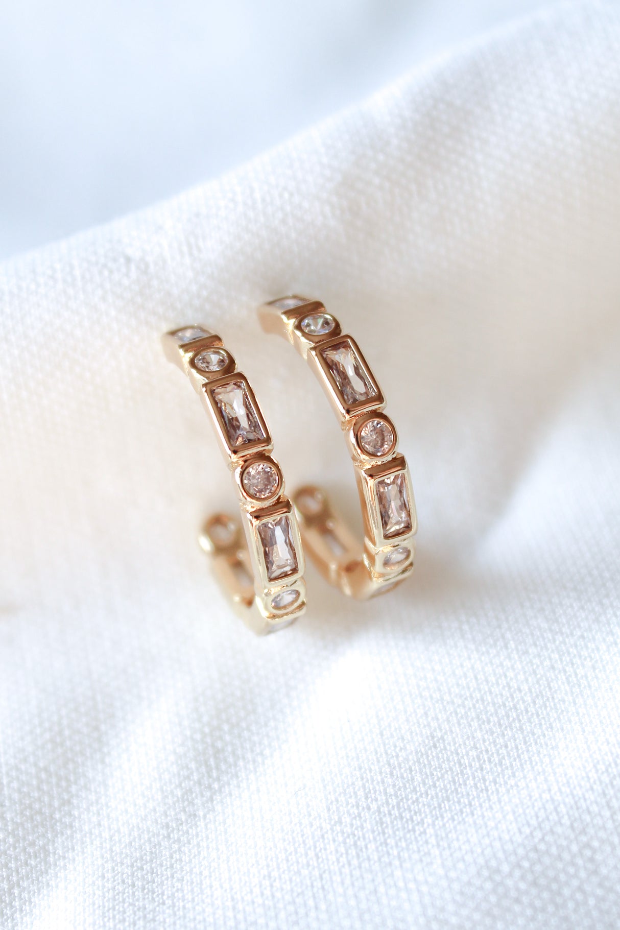 The Ellie Hoop by Kinsey Designs features gold filled hoops with rectangular and round gemstones, shown on a white textured fabric. Discover this elegant jewelry piece at our exclusive sample sale.