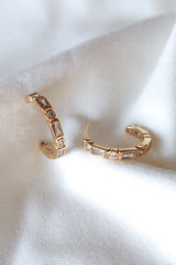 The Ellie Hoop by Kinsey Designs features gold filled hoops with rectangular and round clear stones, displayed on a soft white fabric—an elegant accessory for any occasion.