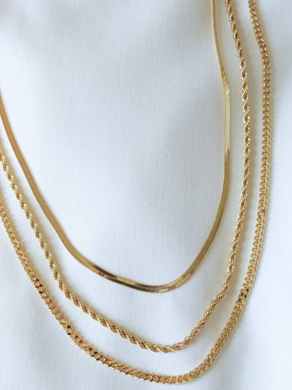 Four gold chains with unique patterns, including the Emile Layer Necklace by Kinsey Designs Archives, are displayed on a white fabric background.