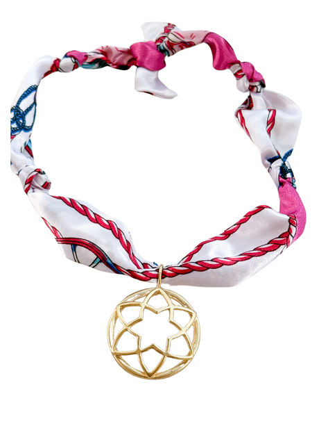 The Florence Scarf Necklace by Kinsey Designs features pink and white patterns, a bold geometric gold pendant, and an adjustable design for the perfect fit.