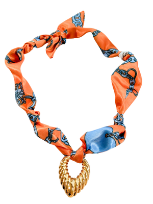 The Como Scarf Necklace by Kinsey Designs features an orange patterned silk scarf with a vintage pendant and twisted gold centerpiece, plus an adjustable design for the perfect fit.