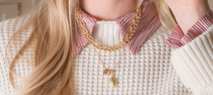 Smiling woman in a cap holding a glass, showing off game-day charm with Kinsey Designs’ Football Grip Charm necklace, paired with layered jewelry, a cream sweater, and a striped shirt.