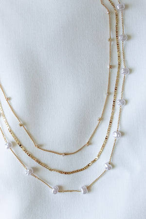 The Folly Layer Necklace by Kinsey Designs Archives features delicate gold chains and small pearls, displayed on a white textured fabric background.