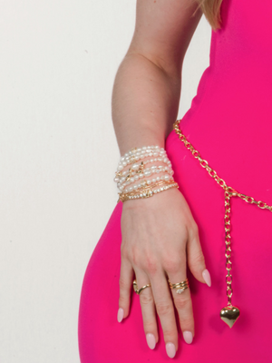 A woman's hand wearing the Kinsey Designs Freshwater Pearl Stretch Bracelet, gold rings, and a bright pink dress with a gold chain belt.
