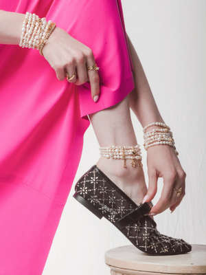 A woman in a bright pink dress, pearl jewelry, and black patterned shoes points to her ankle, showcasing the Kinsey Designs Freshwater Pearl Stretch Anklet.