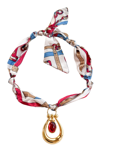 The Genoa Scarf Necklace by Kinsey Designs features a vibrant silk adjustable scarf and a gold vintage pendant with a bold red gemstone—an eye-catching, one-of-a-kind accessory for any wardrobe.