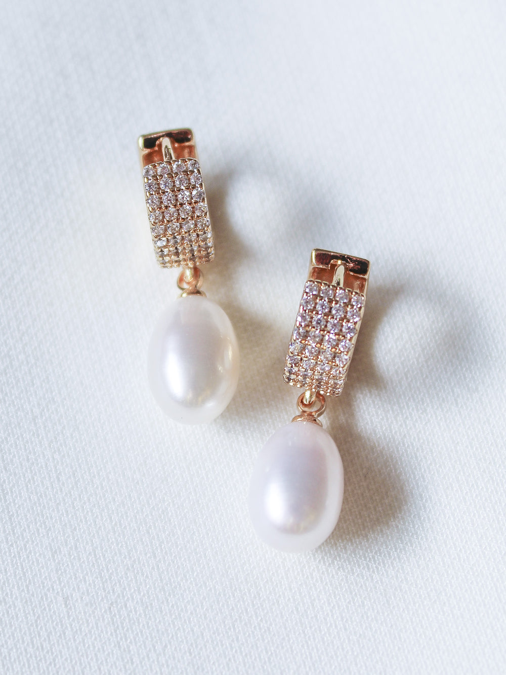 The Gemma Pearl Huggies by Kinsey Designs Archives feature gold-filled hoops with dangling white pearls, elegantly displayed on a white textured fabric background.