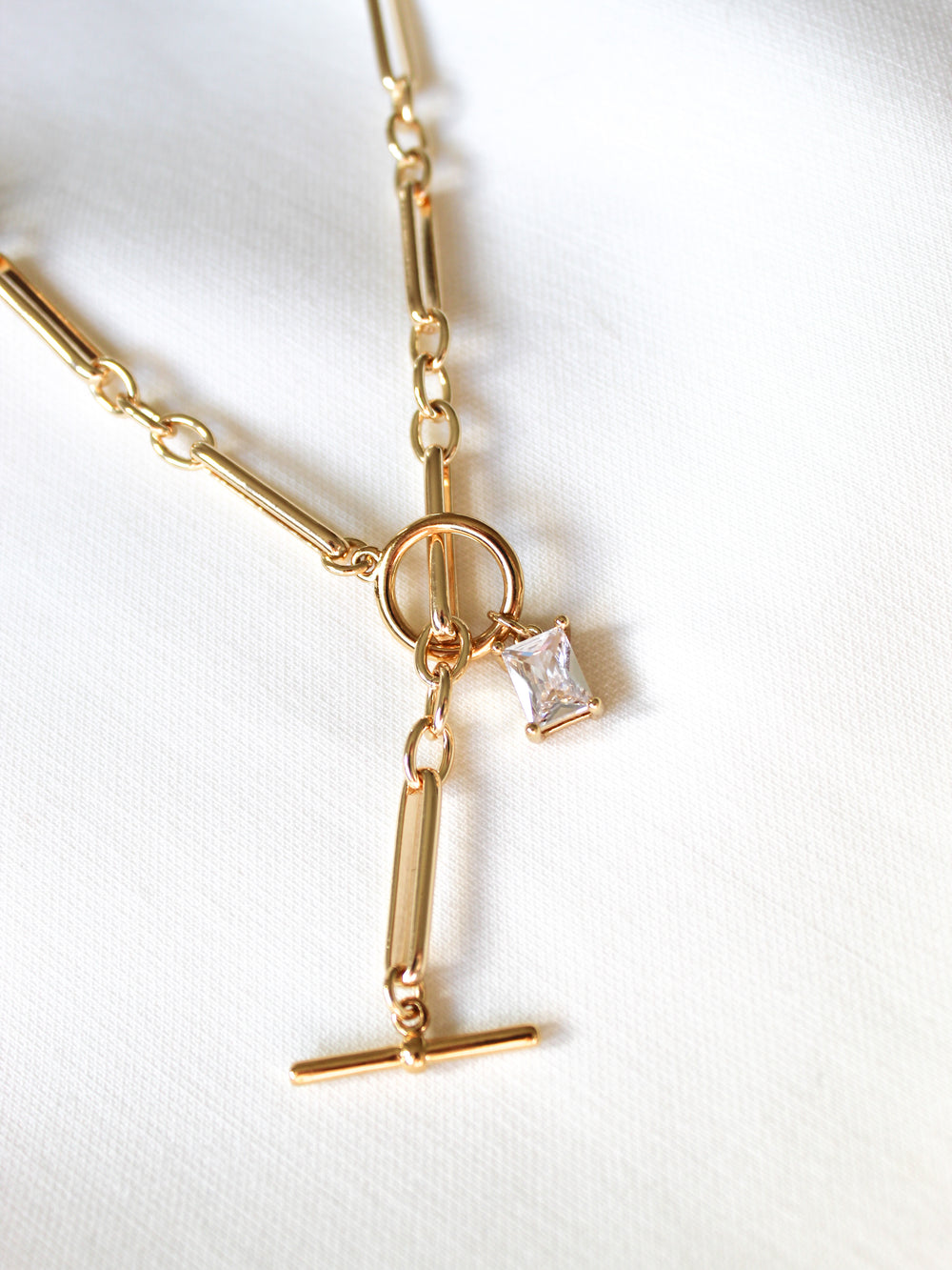 The Kinsey Designs Gilmore Necklace is a gold filled toggle pendant necklace with a rectangular clear gemstone, shown on a white fabric background.