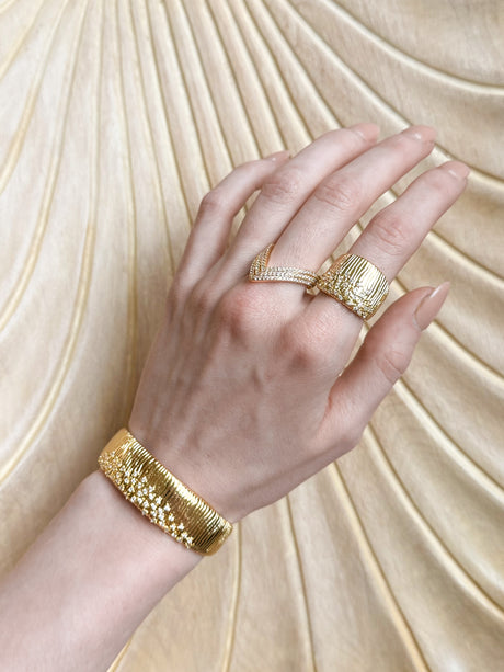 A hand wears gold rings and the Kinsey Designs Glinda Stardust Cuff Bracelet, featuring CZ stones, against a textured cream background.
