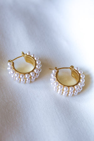 The Hailey Pearl Hoops by Kinsey Designs feature gold hoops adorned with small white pearls, elegantly displayed on white fabric—perfect statement earrings for any occasion.