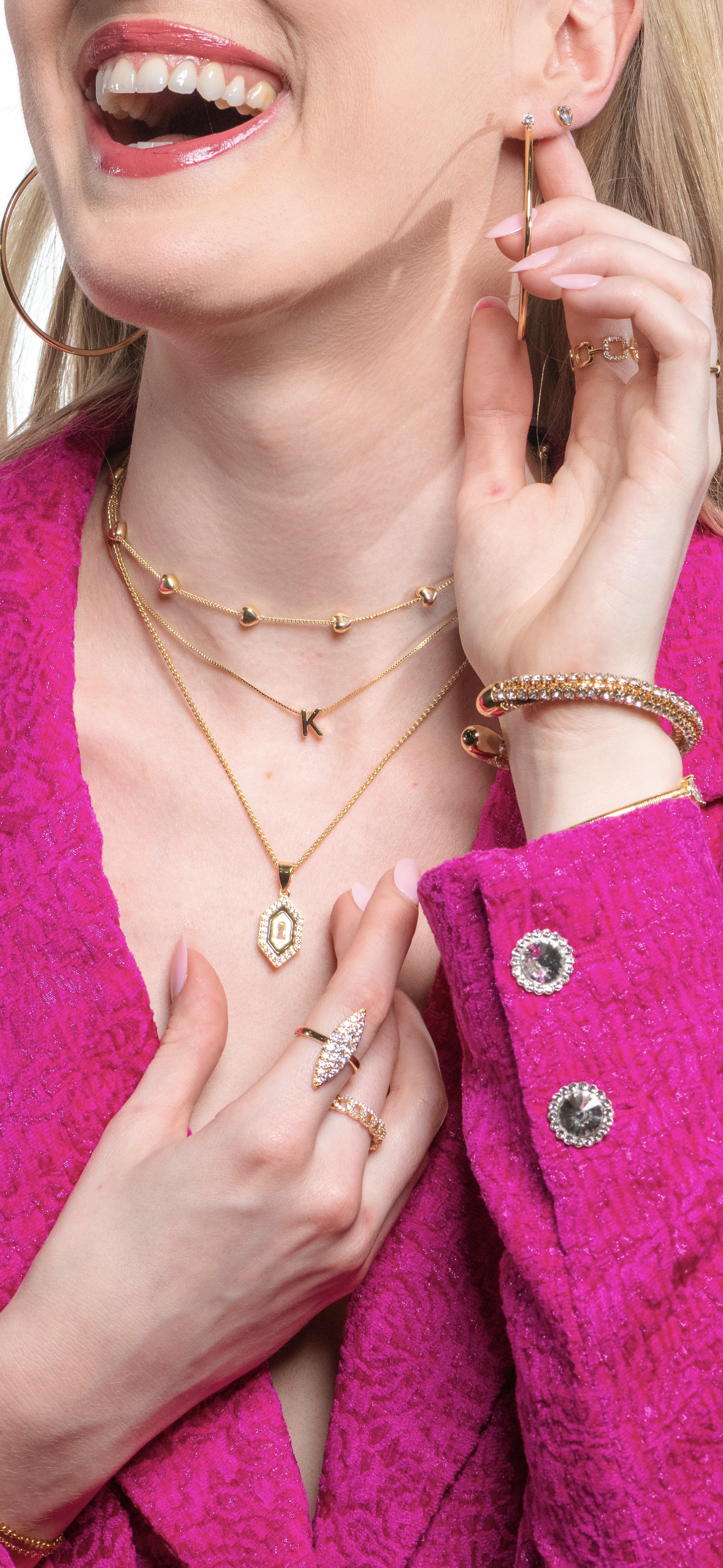 A woman in a pink textured blazer smiles as she touches her earrings and necklaces, layering gold jewelry including the Kinsey Designs Archives Harlowe Necklace—a 17.75-inch chain with a sparkling cubic zirconia pendant.