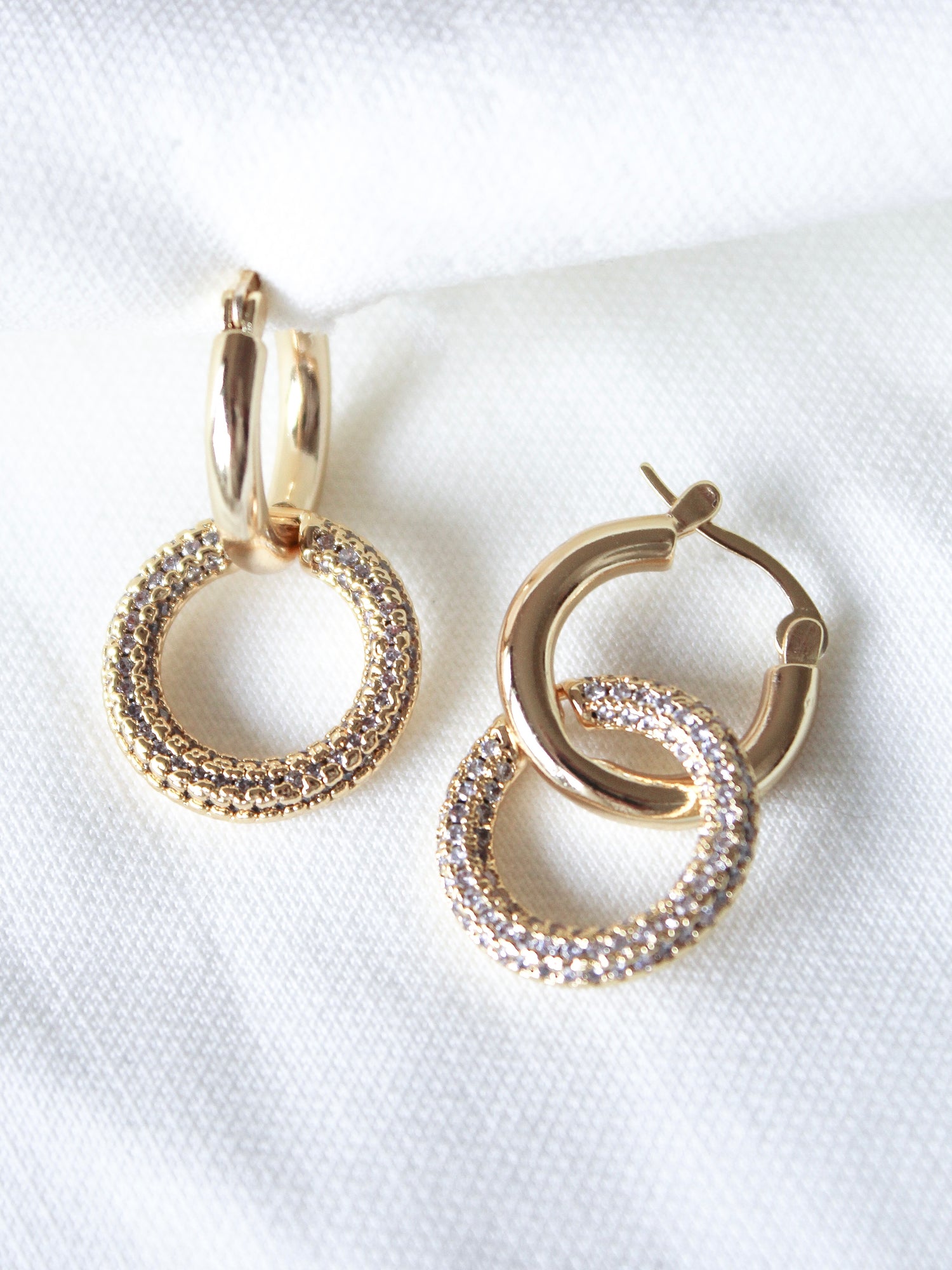 The Holland Hoop by Kinsey Designs features sparkling gold hoops, beautifully displayed on a soft white fabric background.