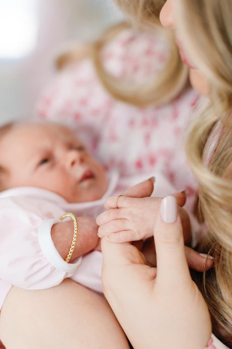 A woman gently holds her newborn’s hand; both wear soft pink clothing, and the mother wears the nickel-free Fallon Mama & Baby Cuff Set by Kinsey Designs.