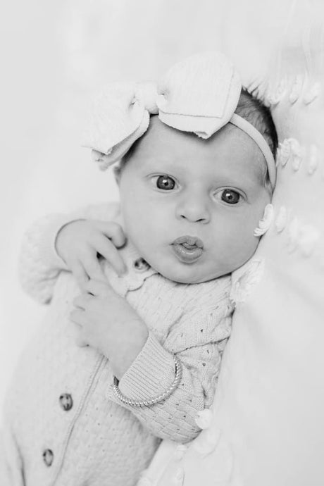 Black and white photo of a baby with a headband and bow, lying down and looking at the camera—just as timeless as the Fallon Baby Cuff by Kinsey Designs.