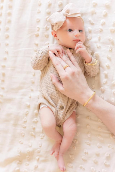A newborn in a beige outfit and bow lies on a textured blanket with an adult hand gently resting on them, showcasing the adjustable Fallon Baby Cuff bracelet by Kinsey Designs.