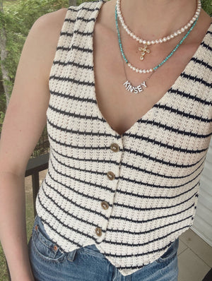 A woman stands on a balcony wearing a striped knit vest, blue jeans, and layered necklaces, including the Bellena Pearl Cross Necklace by Kinsey Designs.