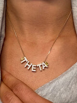 The Kinsey Designs THETA Charm Necklace features diamond-encrusted letters spelling "THETA," elegantly resting on the wearer’s neck.