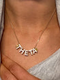 The Kinsey Designs THETA Charm Necklace features diamond-encrusted letters spelling "THETA," elegantly resting on the wearer’s neck.