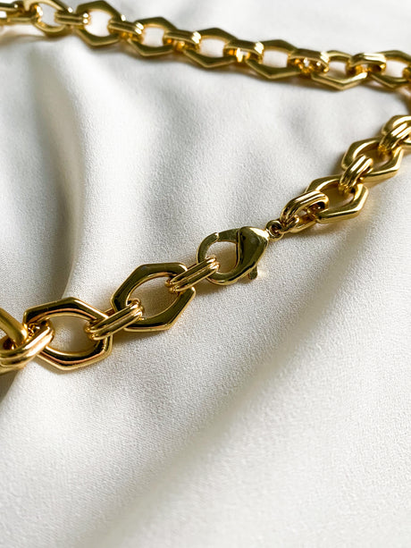 Close-up of the Prague- Slight Flaw gold chain necklace by Kinsey Designs Archives, featuring geometric links on a soft white fabric background.