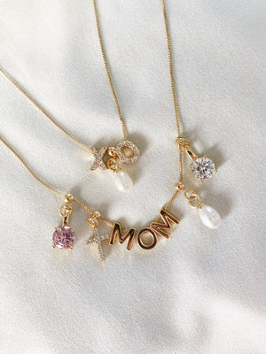 Three gold Kinsey Designs necklaces—charms reading "XO," "MOM," and "S"—feature pearls, gemstones, and the Cross Grip Charm crystal cross, arranged on white fabric.