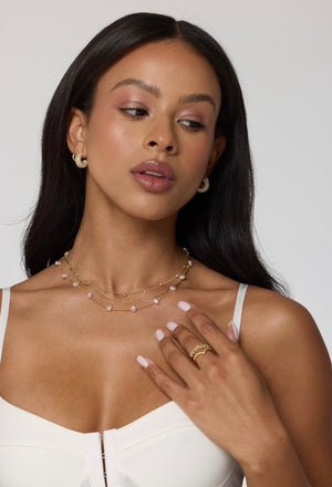 A woman in a white top wears gold hoop earrings, rings, manicured nails, and the Kinsey Designs Archives Folly Layer Necklace—a stylish layered gold chain necklace.