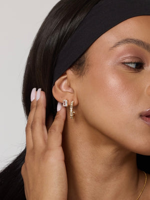 A woman with smooth skin wears Kinsey Designs Prism Studs, a black headband, and light pink nail polish as she touches her ear.
