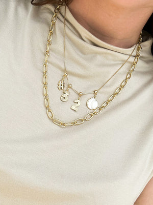 Woman wearing layered gold necklaces, including a Kinsey Designs White Baseball Grip Charm necklace, over a light beige top.