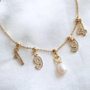 The Kinsey Designs Charm Bracelet Dainty Chain features number charms "1", "9", "9", "4" and a pearl, laid on white fabric—perfect for pairing with your favorite dainty chain or gold-filled bracelet for a chic layered style.